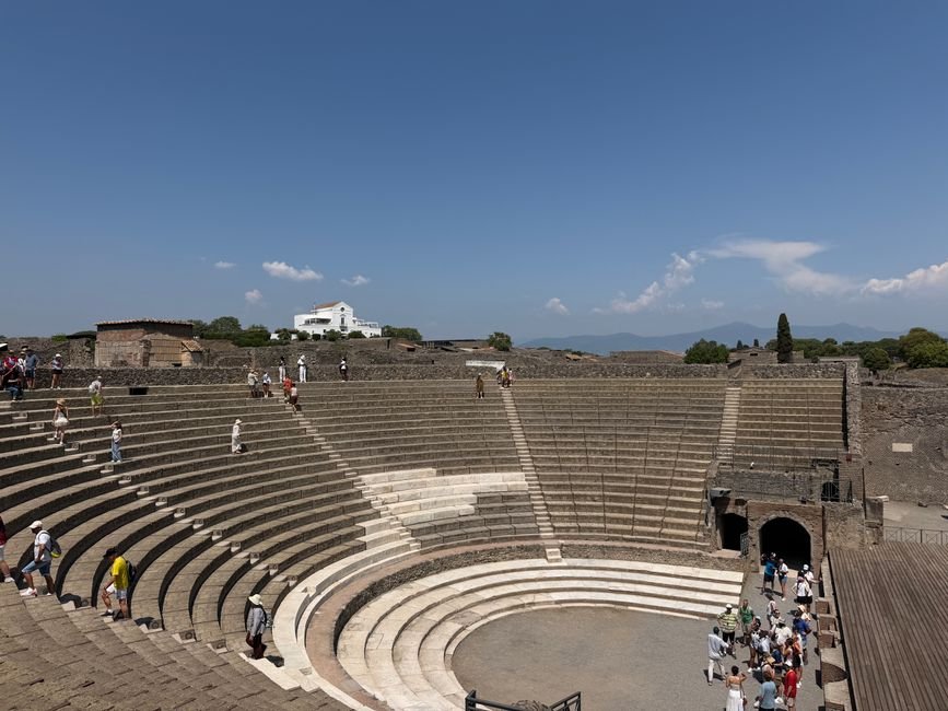 A group of tourists explore the historic amphitheater in Pompeii, Italy, under a clear blue sky.